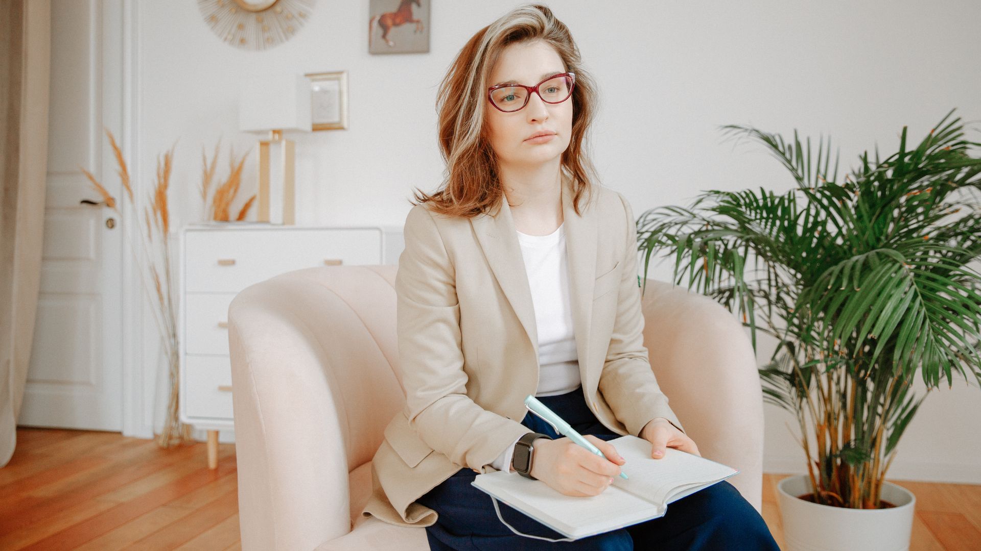 A person with glasses sits on a beige armchair, writing in a notebook. There is a plant and white furniture in the background.